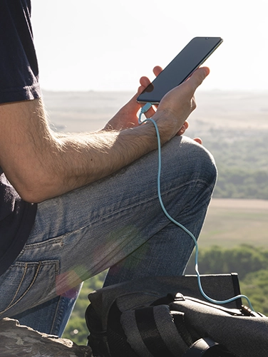Man sits on a hill while hiking and charges his smartphone with the Hama Power Pack "Supreme 20HD"